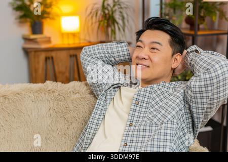 Heureux homme chinois d'âge moyen insouciant assis confortablement avec la main derrière la tête sur le canapé dans le salon à la maison. Souriant mec coréen se relaxant sur le canapé pensant regarder autour dans la chambre de l'appartement. Banque D'Images