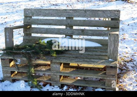 Un banc rustique fabriqué à partir de palettes en bois avec une branche de pin vert et de la neige sur le siège se dresse à l'extérieur sur un sol enneigé, symbolisant la durabilité an Banque D'Images