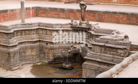 L'ancienne fontaine en pierre présente une sculpture de cobra avec de l'eau qui coule, entourée d'un mur de briques et de détails historiques. Banque D'Images