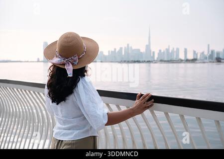 Une femme en chemise blanche et chapeau de paille profite de la vue sur Dubaï avec ses gratte-ciel et ses palmiers Dubai Creek, femme voyageant à Dubaï Émirats arabes Unis Banque D'Images