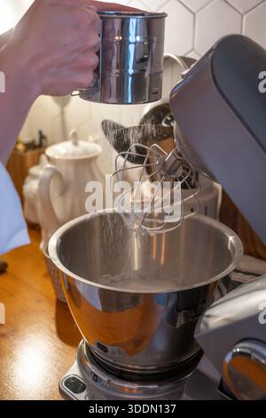 Les mains tamisent la farine dans le bol du mélangeur tout en préparant la pâte charlotte aux pommes sur le comptoir de cuisine en bois, capturant le moment de cuisson à la maison concentré dans une lumière chaude Banque D'Images