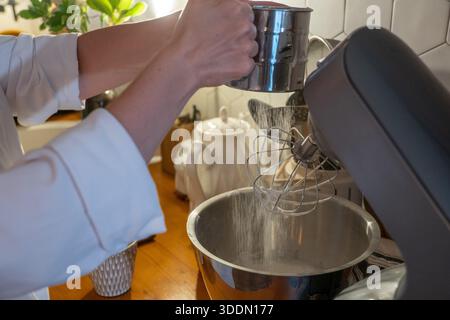 Les mains tamisent la farine dans le bol du mélangeur tout en préparant la pâte charlotte aux pommes sur le comptoir de cuisine en bois, capturant le moment de cuisson à la maison concentré dans une lumière chaude Banque D'Images