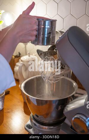 Les mains tamisent la farine dans le bol du mélangeur tout en préparant la pâte charlotte aux pommes sur le comptoir de cuisine en bois, capturant le moment de cuisson à la maison concentré dans une lumière chaude Banque D'Images