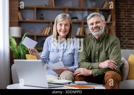 Heureux couple aîné assis sur le canapé à la maison, souriant tout en utilisant un ordinateur portable et en examinant des documents pour calculer les factures et planifier l'épargne-retraite et les finances ensemble Banque D'Images