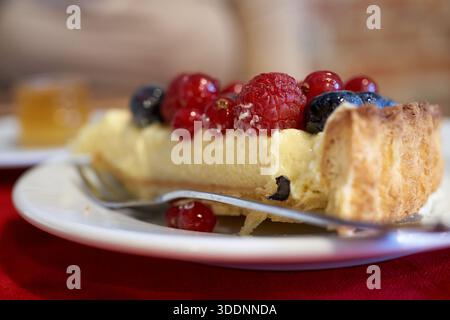 Gros plan d'une tranche de cheesecake garnie de framboises, de groseilles rouges et de myrtilles sur une assiette blanche, cadre dessert de café. Banque D'Images
