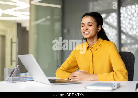 Jeune femme d'affaires avec les yeux fermés souriant, trouvant un moment de pleine conscience et de détente pendant sa journée de travail, prenant une pause de l'ordinateur portable dans un environnement de bureau moderne Banque D'Images