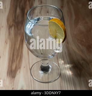 Eau plate pure dans un verre avec une tranche de citron jaune, placée sur une table en bois clair Banque D'Images