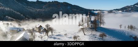 Vue panoramique de l'église Jamnik et du village dans le brouillard d'hiver : Saint Primus et l'église Felician sur une colline enneigée au lever du soleil, Alpes slovènes. Banque D'Images