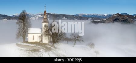 Vue panoramique de l'église Jamnik et du village dans le brouillard d'hiver : Saint Primus et l'église Felician sur une colline enneigée au lever du soleil, Alpes slovènes. Banque D'Images