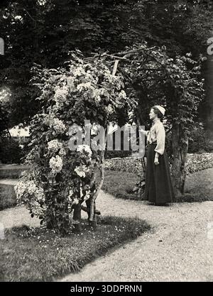 Une femme posant à une arche en bois rustique (arche, tonnelle ou tonnelle) avec des roses en fleurs au Royaume-Uni vers 1900. Le look rustique était très populaire parmi les jardiniers victoriens. C’était une ère de grands changements – avec le travail désormais centré dans les villes industrielles, la main-d’œuvre du pays s’est éloignée d’un passé agricole. Le look rustique reflétait ces simples campagnes daysrurales – une photographie vintage des années 1800/1900. Banque D'Images
