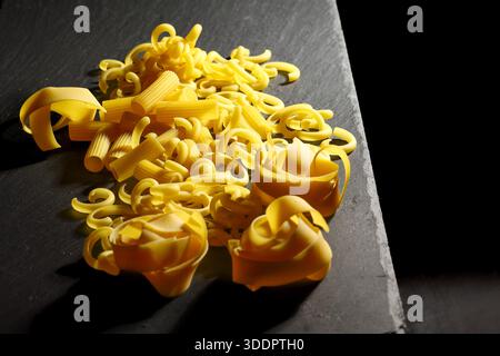 Vue rapprochée d'un plat de pâtes crues italiennes traditionnelles de différentes formes et types, préparées sur un plateau en pierre sombre pour la préparation et la cuisson d'un Itali Banque D'Images