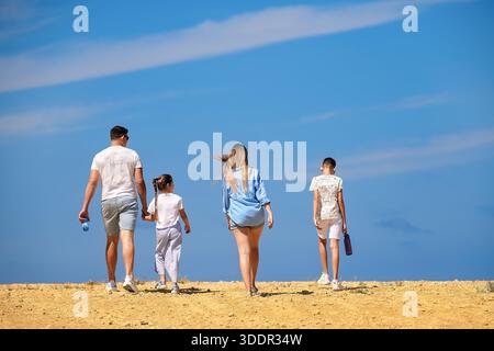 Une famille de quatre personnes en tenues blanches et bleues marche à travers un désert rocheux chaud et sec contre un ciel bleu, père tenant la main de sa fille. Banque D'Images
