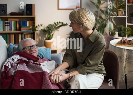 Homme caucasien senior allongé sur un canapé couvert d'une couverture, se tenant la main avec une femme caucasienne d'âge moyen assise à côté de lui, montrant soutien et soins dans le cadre familial Banque D'Images