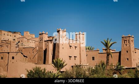 Vue rapprochée des bâtiments de forteresse en terre à ait Ben Haddou, Maroc, avec de hautes tours, des palmiers et un ciel bleu clair en plein jour. Banque D'Images