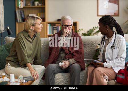 Homme caucasien senior assis sur un canapé toussant tout en tenant des médicaments, femme caucasienne d'âge moyen le soutenant, jeune adulte médecin féminin noir consultant et prenant des notes Banque D'Images