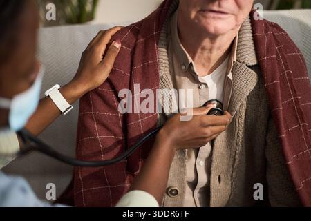 Homme caucasien senior assis tandis que femme noire travailleur de la santé utilisant le stéthoscope pour écouter la poitrine, soignant plaçant doucement la main sur l'épaule des patients, fermer l'examen médical Banque D'Images