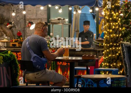 Sunset Town sur l'île de Phu Quoc. 23 décembre 2025. Homme ayant une bière artisanale premium dans un comptoir de bar du soir avec des décorations festives et un chef Banque D'Images