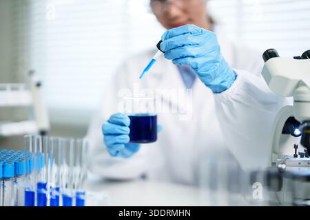Scientifique adulte diligent effectue des analyses chimiques précises dans un laboratoire moderne. Mains porter des gants de protection transférant la solution bleu vif pendant Banque D'Images