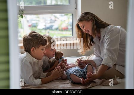 Mère et deux enfants jouent avec un nouveau-né sur un lit près d'une fenêtre, lumière du jour douce, couleurs neutres, atmosphère familiale chaleureuse et attentionnée Banque D'Images