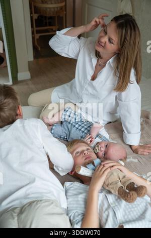 La mère et les enfants sont allongés sur le sol avec un nouveau-né à la maison, une lumière du jour douce, des couleurs neutres, une interaction familiale calme et intime Banque D'Images
