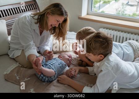Mère et deux enfants jouent avec un nouveau-né sur un lit près d'une fenêtre, lumière du jour douce, couleurs neutres, atmosphère familiale chaleureuse et attentionnée Banque D'Images