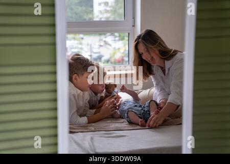 Mère et deux enfants jouent avec un nouveau-né sur un lit près d'une fenêtre, lumière du jour douce, couleurs neutres, atmosphère familiale chaleureuse et attentionnée Banque D'Images