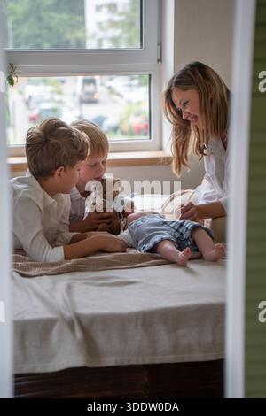Mère et deux enfants jouent avec un nouveau-né sur un lit près d'une fenêtre, lumière du jour douce, couleurs neutres, atmosphère familiale chaleureuse et attentionnée Banque D'Images