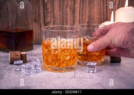 Une main tient un gobelet à whisky à côté d'un autre verre sur de la glace, avec une bouteille et des bougies en arrière-plan. Concept de bar Moody pour partager des boissons, alcool nous Banque D'Images
