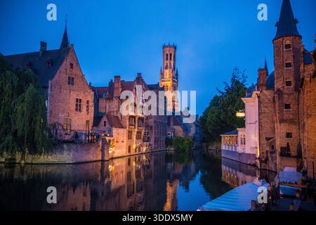 Crépuscule à Bruges : le beffroi et les bâtiments médiévaux illuminés le long du canal de Dijver, encadrés par un arbre silhouette dans le cœur historique de la Belgique. Banque D'Images