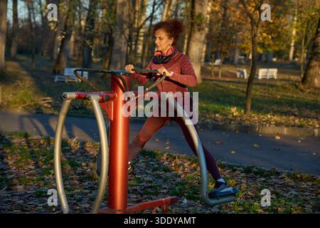 La femme athlétique exerce sur le cross trainer elliptique au gymnase extérieur dans le parc d'automne avec des feuilles mortes Banque D'Images