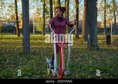 La femme athlétique exerce sur le cross trainer elliptique au gymnase extérieur dans le parc d'automne avec des feuilles mortes Banque D'Images
