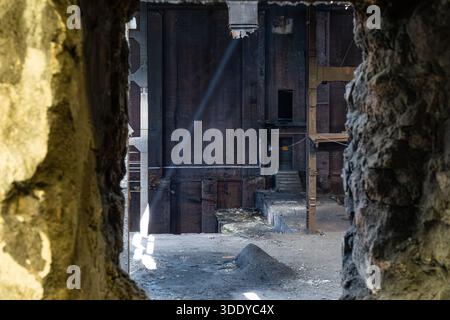 Vue intérieure d'un bâtiment industriel abandonné vu à travers un mur de béton endommagé avec des structures en acier et de la lumière naturelle Banque D'Images