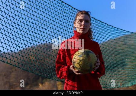 Femelle en tricot rouge tient le volley jaune, encadré par un filet vert avec toile de fond montagneuse, couleurs d'automne, et l'eau coulante à proximité. Banque D'Images