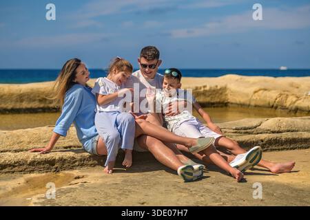Famille heureuse avec deux enfants se détend sur les rochers au bord de la mer : fille et fils jouent avec papa, tandis que maman les regarde souriants. Banque D'Images