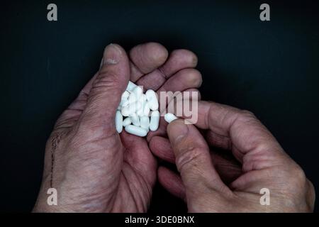 Gros plan de mains masculines tenant des pilules blanches, avec une pilule prise, isolée sur un fond noir, symbolisant un médicament ou une consommation de drogue. Banque D'Images
