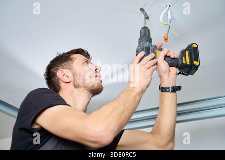 L'homme portant une combinaison de travail perce un trou dans le plafond à l'aide d'une perceuse électrique sans fil dans la pièce intérieure. Concept de rénovation domiciliaire, d'installation et de service d'entretien professionnel. Banque D'Images