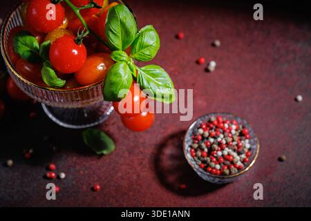 Vue de dessus de tomates cerises avec basilic dans un bol en verre et poivre mélangé sur une table rouge Banque D'Images