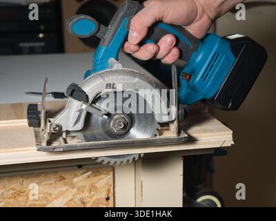 Main utilisant une scie circulaire sans fil pour couper du contreplaqué ; gros plan de la main d'un ouvrier tenant et utilisant une scie circulaire alimentée par batterie pour couper un plan en bois Banque D'Images