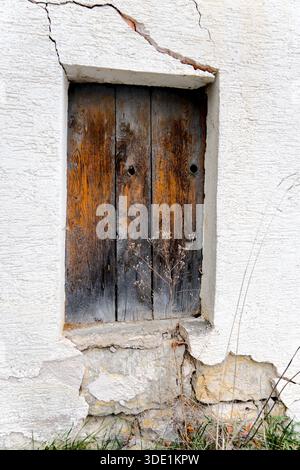 Fenêtre en bois abandonnée placée dans un mur de plâtre fissuré, entourée de mauvaises herbes envahies et de pourriture naturelle. Banque D'Images
