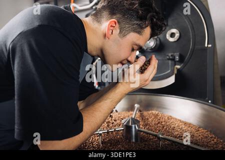 Vue latérale du jeune travailleur sentant des grains de café torréfiés frais au-dessus du tambour de la machine vue latérale du jeune travailleur sentant des grains de café torréfiés frais abo Banque D'Images