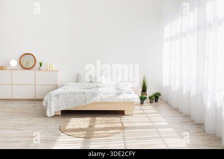 Lit, tapis sur plancher en bois dans la lumière du matin de la fenêtre avec rideaux, table, plantes, meubles Banque D'Images
