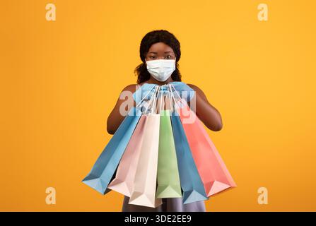 Femme noire millénaire portant un masque facial et des gants en caoutchouc, montrant des sacs-cadeaux à l'appareil photo, faisant des achats pendant le covid Banque D'Images