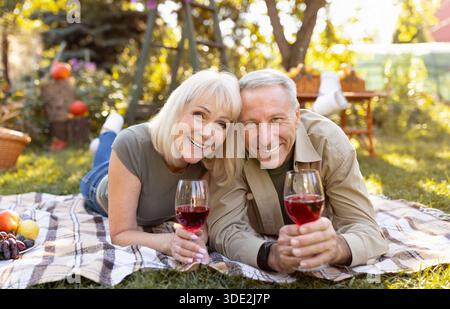 Portrait de couple senior aimant buvant du vin à l'extérieur, allongé sur une couverture tout en pique-niquant dans le jardin Banque D'Images