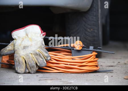 Une paire de gants de travail usés repose sur une rallonge orange vif soigneusement enroulée avec une bobine noire et une fiche orange, positionnée sur une surface en béton Banque D'Images