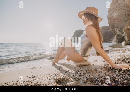 Une jeune femme en maillot de bain argenté métallique et chapeau de paille est assise sur un rocher à une plage de galets, regardant l'océan calme pendant une journée ensoleillée. Banque D'Images