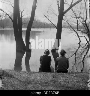 Une photographie d'archive monochrome des années 1980 montre deux femmes assises sur la berge herbeuse, regardant l'eau de fond inondée de la rivière Seversky Donets près de Brusino, région de Donetsk, Ukraine URSS. Les arbres se tiennent submergés dans les hautes eaux de source, projetant des reflets. Ce moment serein et franc capture les résidents locaux contemplant les inondations saisonnières du paysage pendant une période paisible dans le Donbass, reflétant la vie quotidienne et le lien avec la nature avant la guerre. Banque D'Images