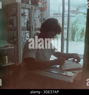 Crimée, URSS, années 1960 Une authentique photographie d'archives en couleur vintage prise à l'intérieur du camp des jeunes pionniers d'Artek. Une travailleuse médicale (médecin ou infirmière) vêtue d'un manteau d'uniforme blanc s'assoit à un bureau, écrivant des notes dans un journal. Derrière elle, une armoire en verre contenant des bouteilles médicales et des fournitures est visible. Une grande fenêtre donne sur le parc verdoyant à l'extérieur. L'image capture l'atmosphère professionnelle, les soins de santé et la routine quotidienne du personnel dans le célèbre camp de jeunes soviétique. Banque D'Images