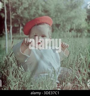 Slaviansk, région de Donetsk, Ukraine, URSS, début des années 1960 Une captivante photographie d'archives en couleur vintage d'une jolie petite fille, Inessa Mikhailovna Kravelis, assise dans de hautes herbes dans un parc ensoleillé. L'enfant porte une tenue d'été blanc clair et un béret rouge vif. Elle atteint sa main vers la caméra, tenant un brin d'herbe, regardant le spectateur avec une expression focalisée. L'arrière-plan présente des arbres verts. Cette image authentique capture l'innocence, la curiosité et l'atmosphère heureuse de l'enfance dans le Donbass paisible à l'époque soviétique. Banque D'Images