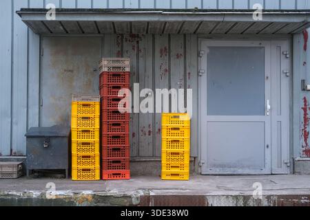 Caisses en plastique jaune et rouge empilées sur un sol en béton sous un auvent métallique, flanquées de deux portes métalliques serties dans un mur industriel gris. Banque D'Images