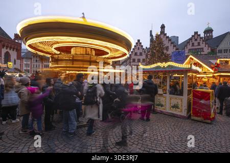 22 décembre 2025, marché de Noël de Francfort sur Roemerberg avec la ligne d'horizon en arrière-plan au crépuscule. Les lumières brillent et les enfants sont carous Banque D'Images